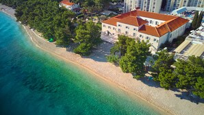 An aerial view of a coastal hotel with a beach, clear waters, and surrounding greenery. - Heritage Hotel Kaštelet (Tucepi)