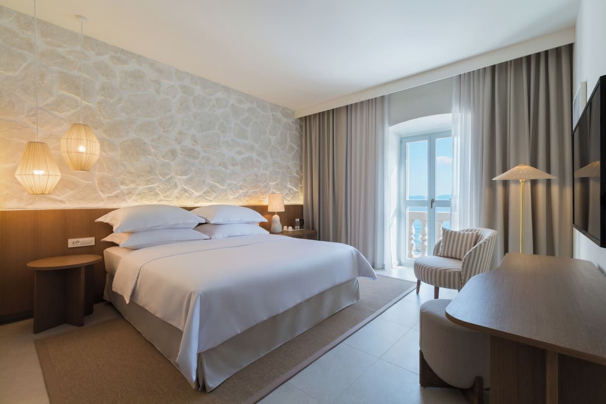 Et moderne hotelværelse med en stenvæg, en stor seng med hvide sengetøj, et siddeområde med et træbord og stole, samt en balkon med gardiner.