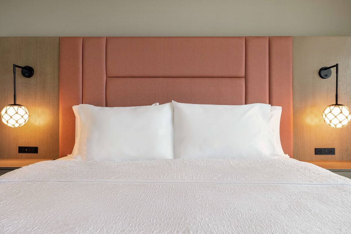 A neatly made bed with a textured white duvet, a padded headboard, and two wall-mounted lamps with geometric light fixtures.
