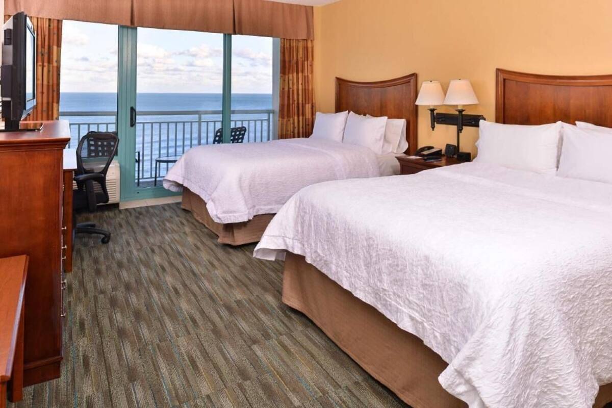 A hotel room with two beds, a desk, a chair, a nightstand with lamps, and a view of the ocean.