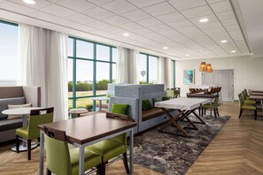 A modern hotel lobby with a seating area, a dining table, and large windows offering a view of the outdoors.