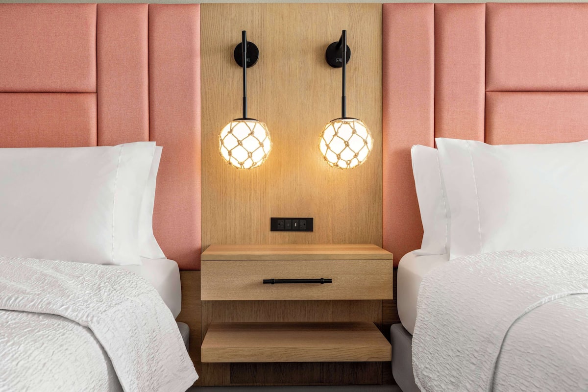 A hotel room with two beds, a wooden nightstand with a drawer, and two wall-mounted lamps.