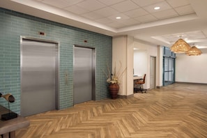 A modern lobby with two elevators, a wooden herringbone floor, a potted plant, and a hanging light fixture.