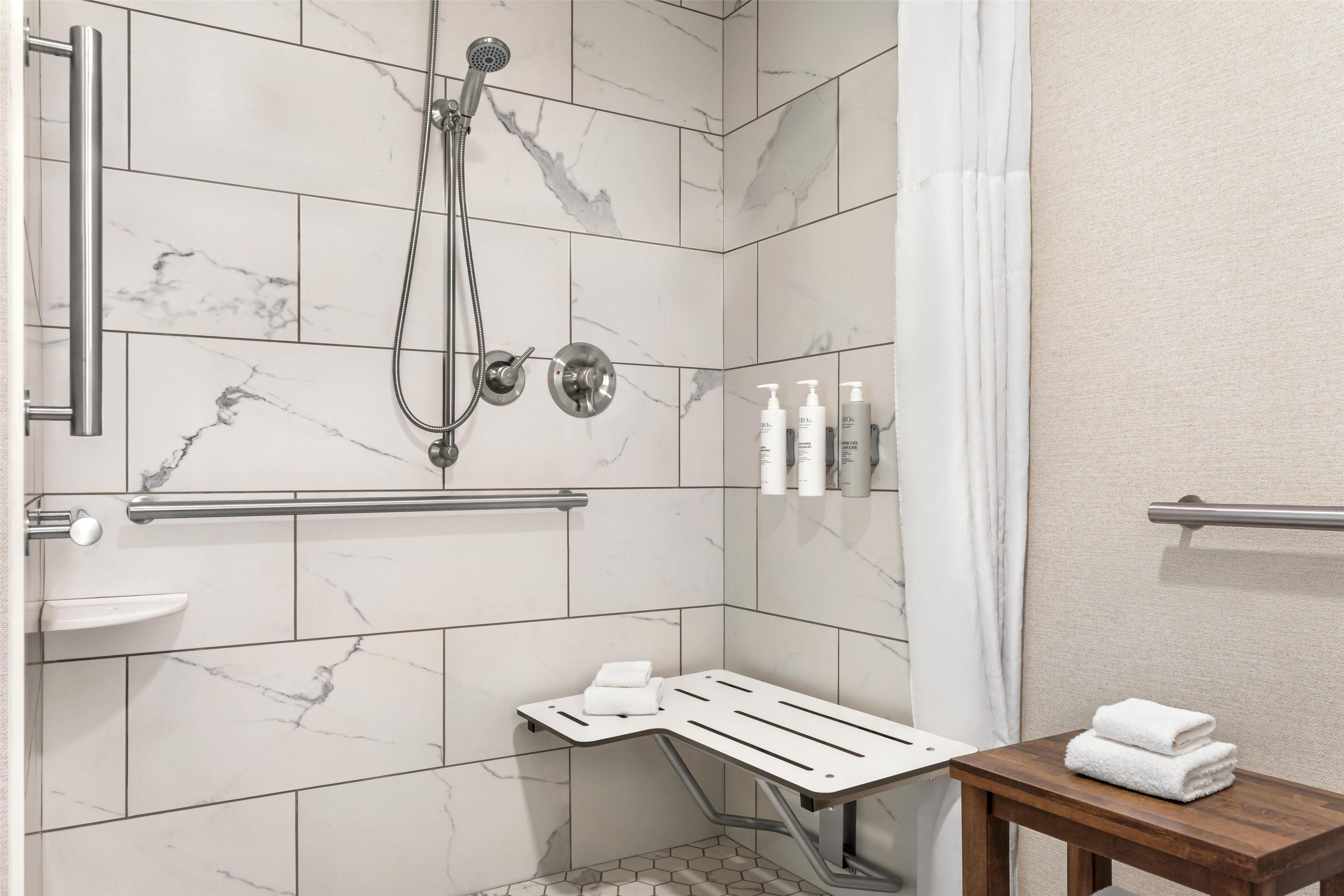 Een moderne badkamer met marmeren tegels, een douche met handdouche, een handgreep, een houten bankje en een handdoekhouder.