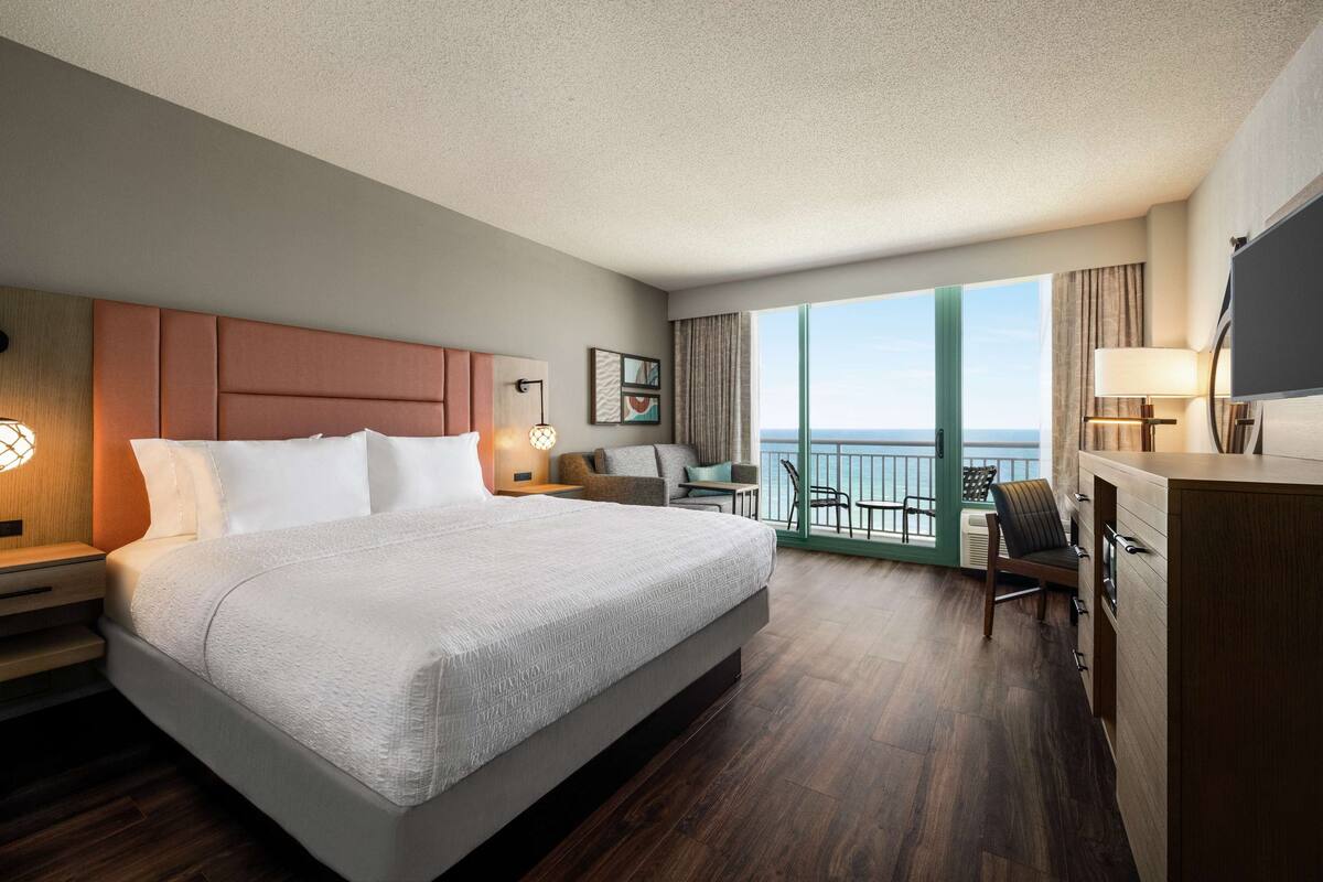 A hotel room with a large bed, a desk, a chair, and a balcony with a view of the ocean.