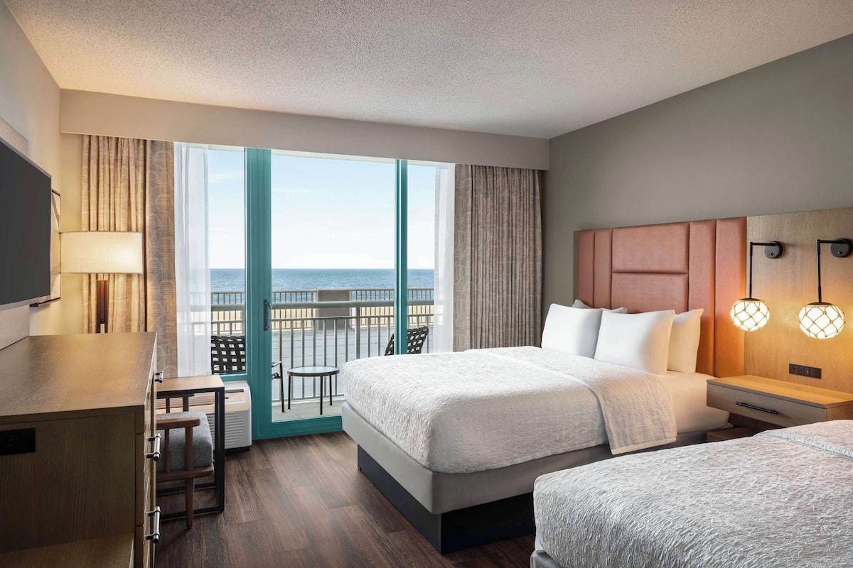 A hotel room with a bed, bedside tables, a desk, a chair, and a view of the ocean.