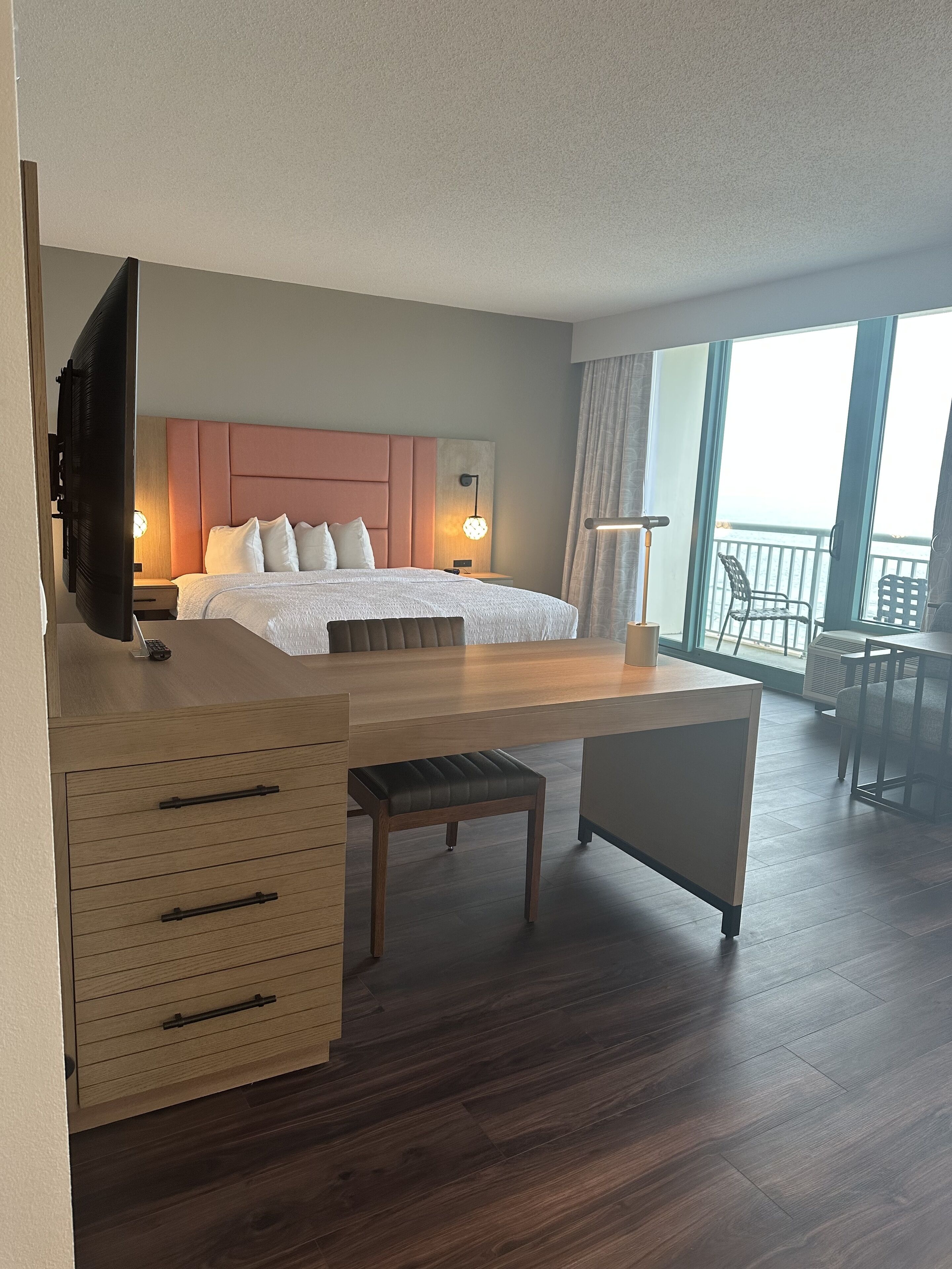 Ein Hotelzimmer mit Bett, Nachttisch, Schreibtisch und Stuhl. Ein Fernseher ist an der Wand montiert, ein Fenster bietet einen Ausblick und ein Balkon lädt mit Sitzgelegenheiten zum Verweilen ein.