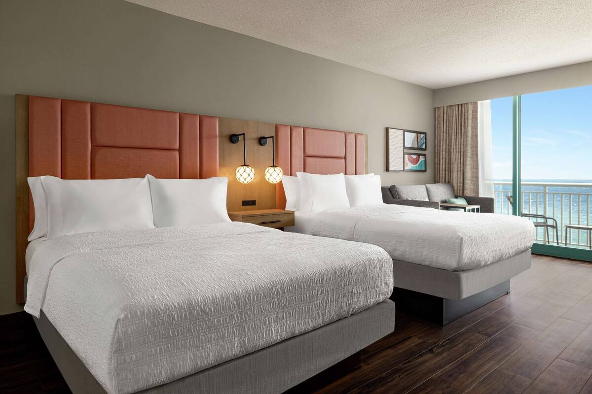 A hotel room with two beds, a nightstand, a desk, and a view of the ocean.