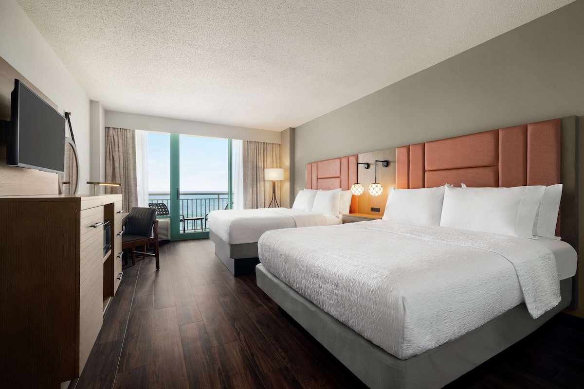 A hotel room with two beds, a desk, a TV, and a balcony with curtains.