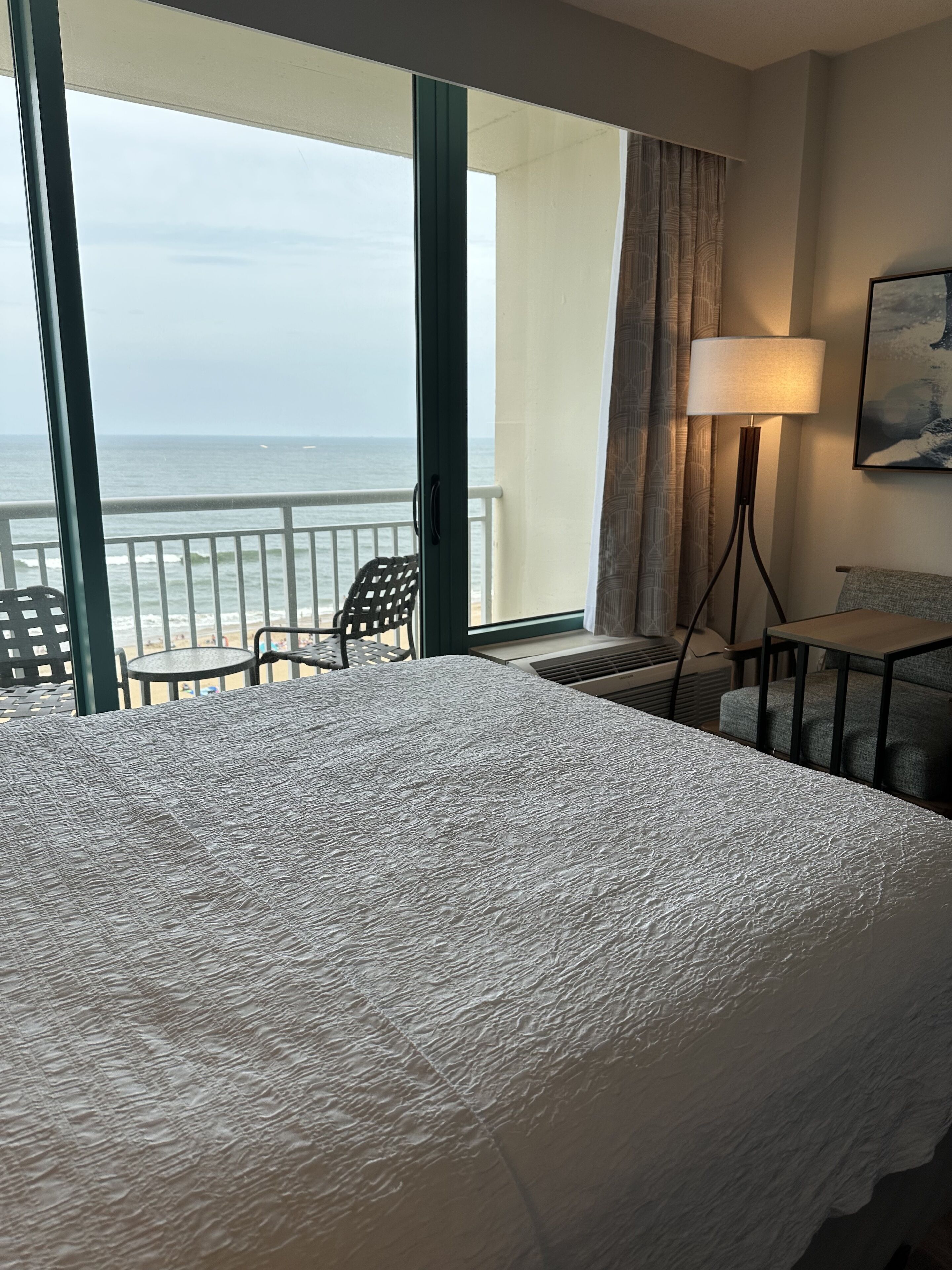 A hotel room with a large bed, a small table, a chair, a lamp, and a view of the ocean.