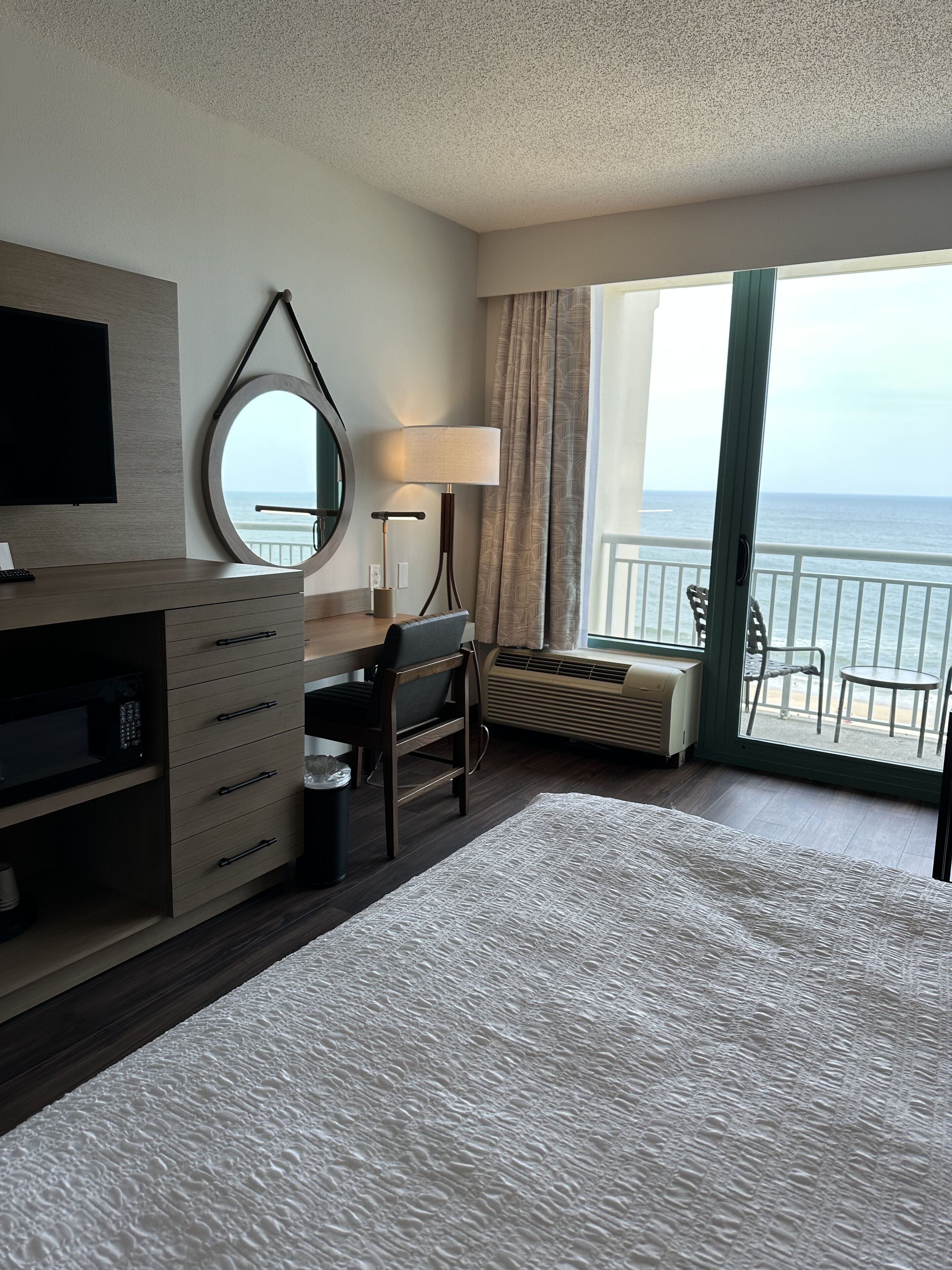 A hotel room with a bed, a desk, a chair, a TV, a lamp, a window with a view of the ocean, and a balcony with chairs.