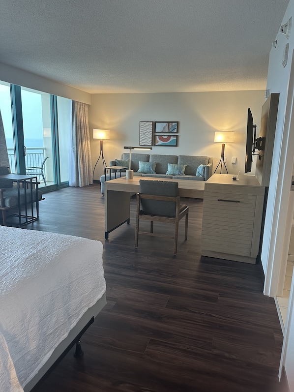 A hotel room with a bed, a desk, a chair, a sofa, a TV, and a balcony with curtains.