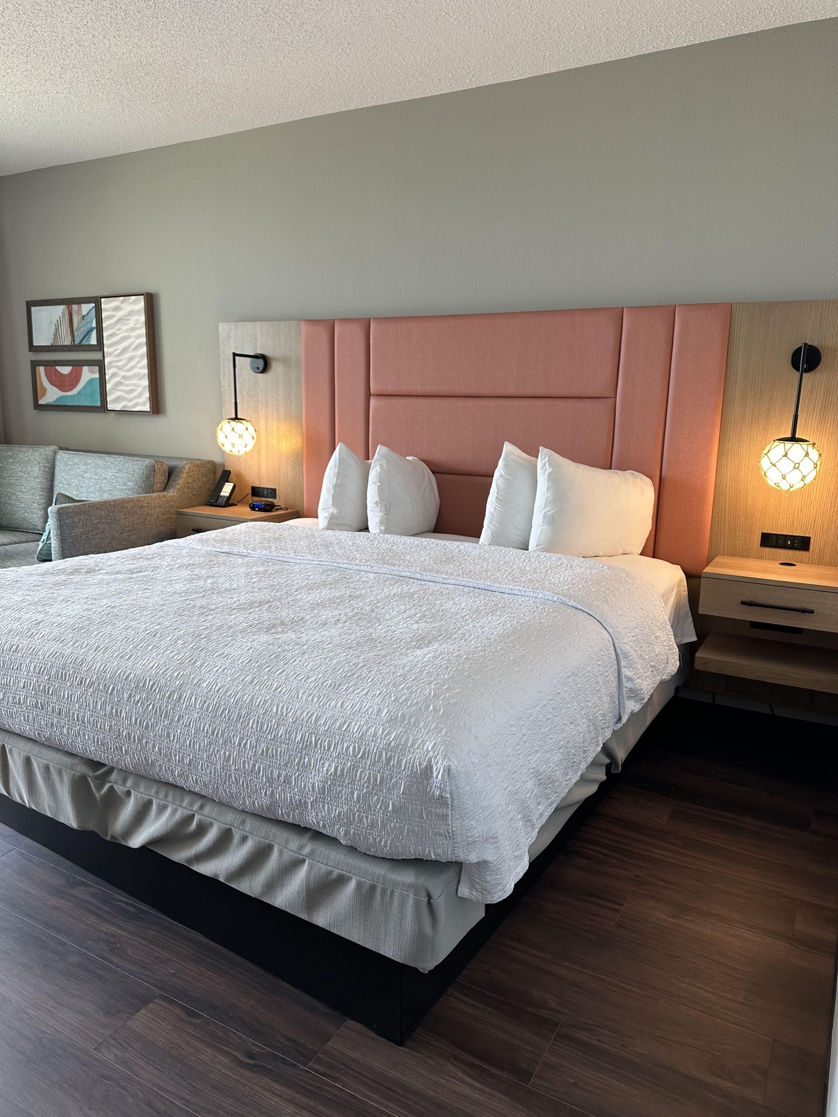 Una habitación de hotel moderna con una cama grande, cabecero, mesitas de noche, una lámpara y una obra de arte en la pared.
