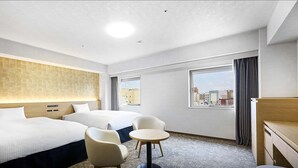 Quarto de hotel com duas camas, uma mesa redonda pequena, duas cadeiras, uma escrivaninha e vista para a cidade pela janela.