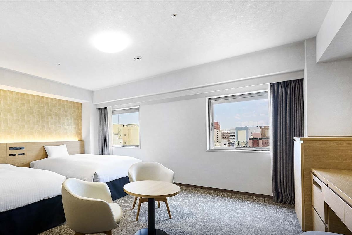 A hotel room with two beds, a small round table, two chairs, a desk, and a view of the city through a window.