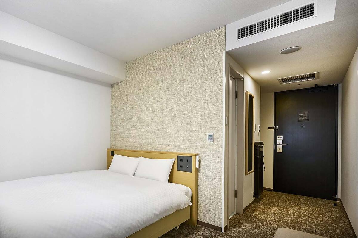 A hotel room with a bed, a desk, and a chair. There is a door and a wall with a textured pattern.
