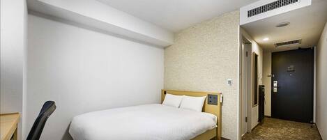 A hotel room with a bed, a desk, and a chair. There is a door and a wall with a textured pattern.