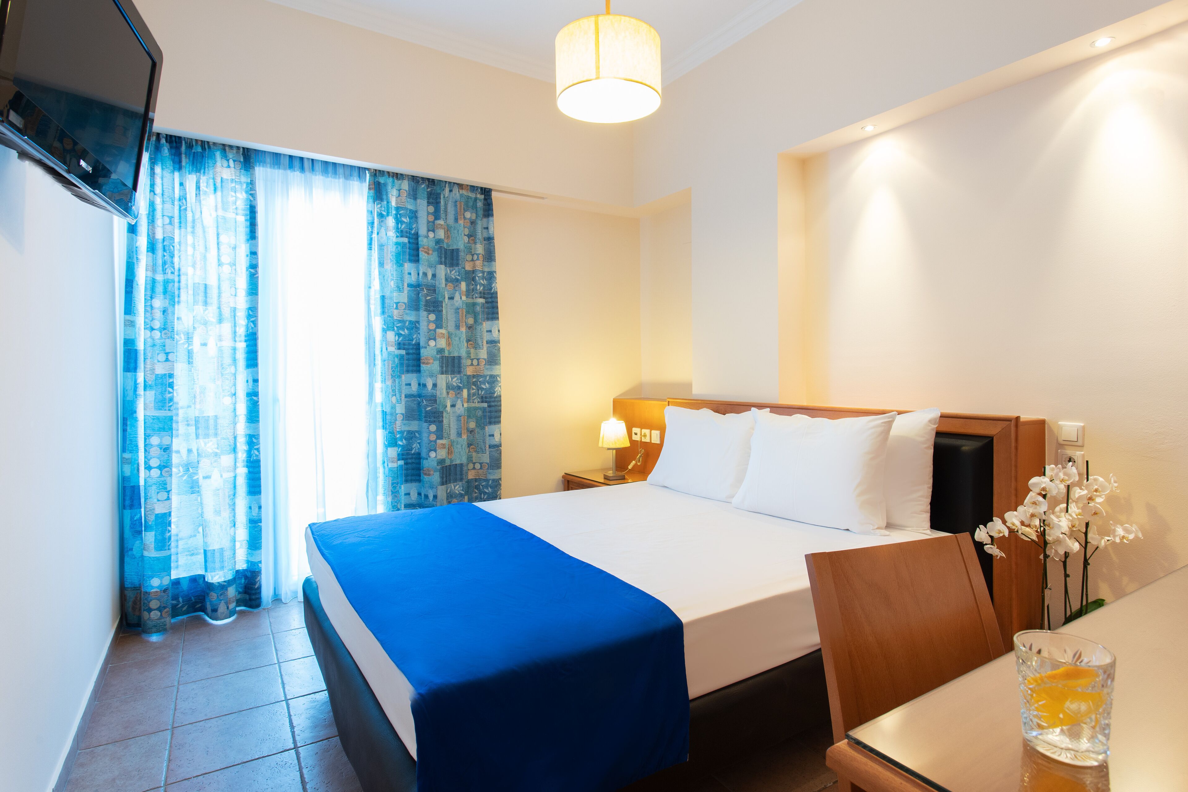 Ein Hotelzimmer mit einem großen Bett, einem Nachttisch mit Lampe, einem an der Wand montierten Fernseher und einem Fenster mit gemusterten Vorhängen.
