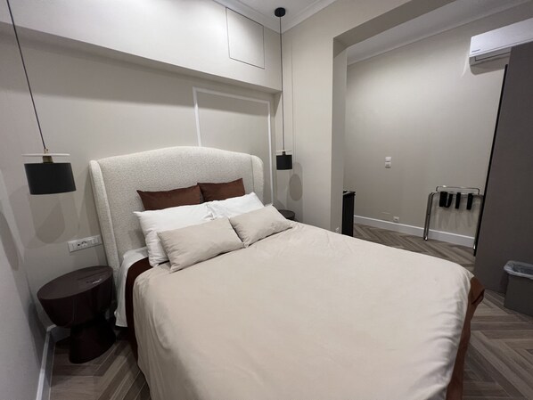 A modern bedroom with a large bed, a bedside table, a wall-mounted lamp, and a door leading to another room. - Pantheon City Hotel (East Mani)