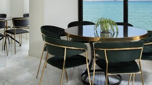 Uma sala de jantar com uma mesa redonda e cadeiras, um vaso com planta e vista para o mar.
