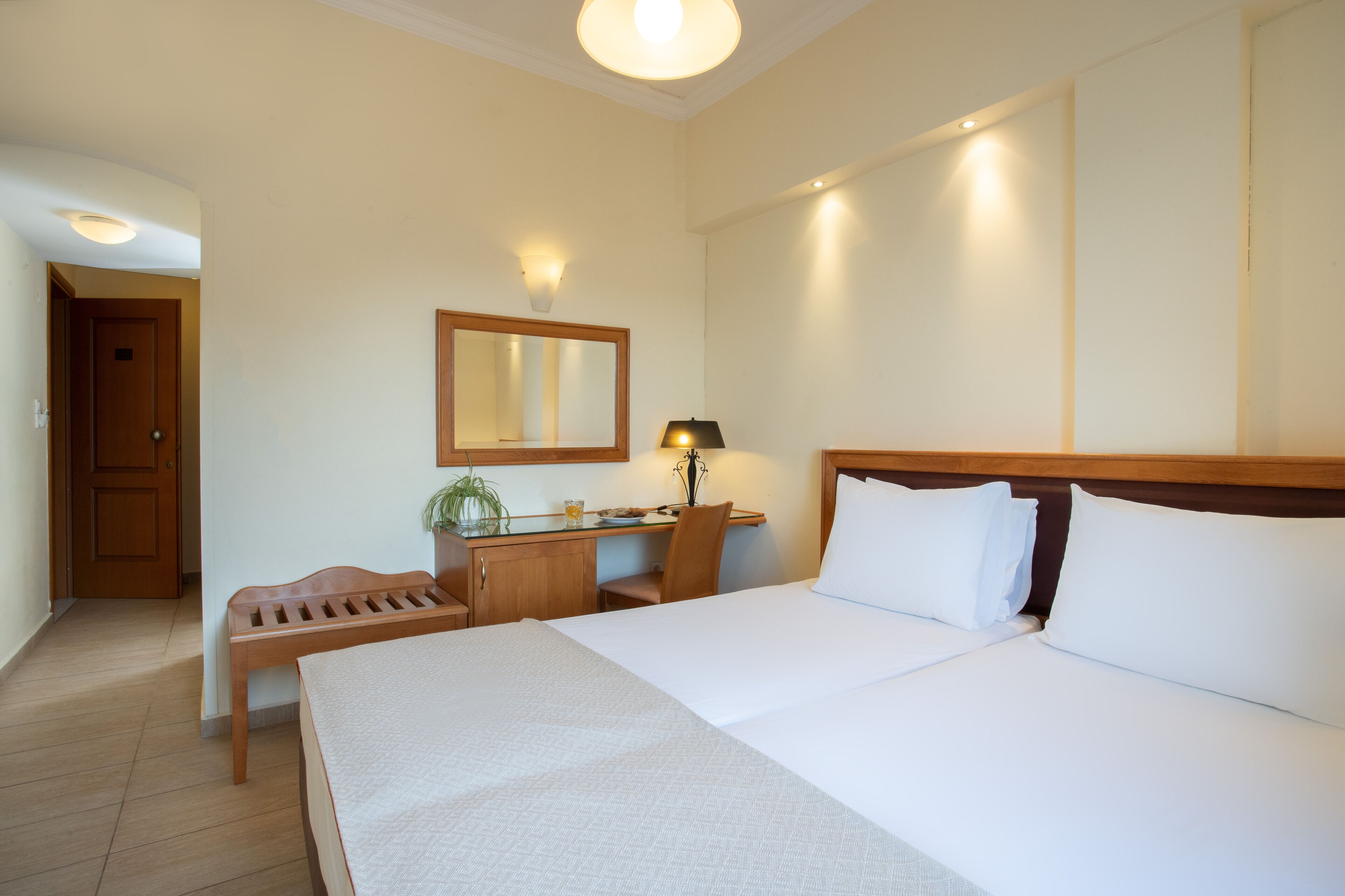 A hotel room with a large bed, a desk with a chair, a small table, a mirror, and a bench.