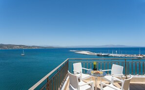 Un balcón con muebles de mimbre blanco que da a un puerto deportivo con barcos y un mar azul cristalino.