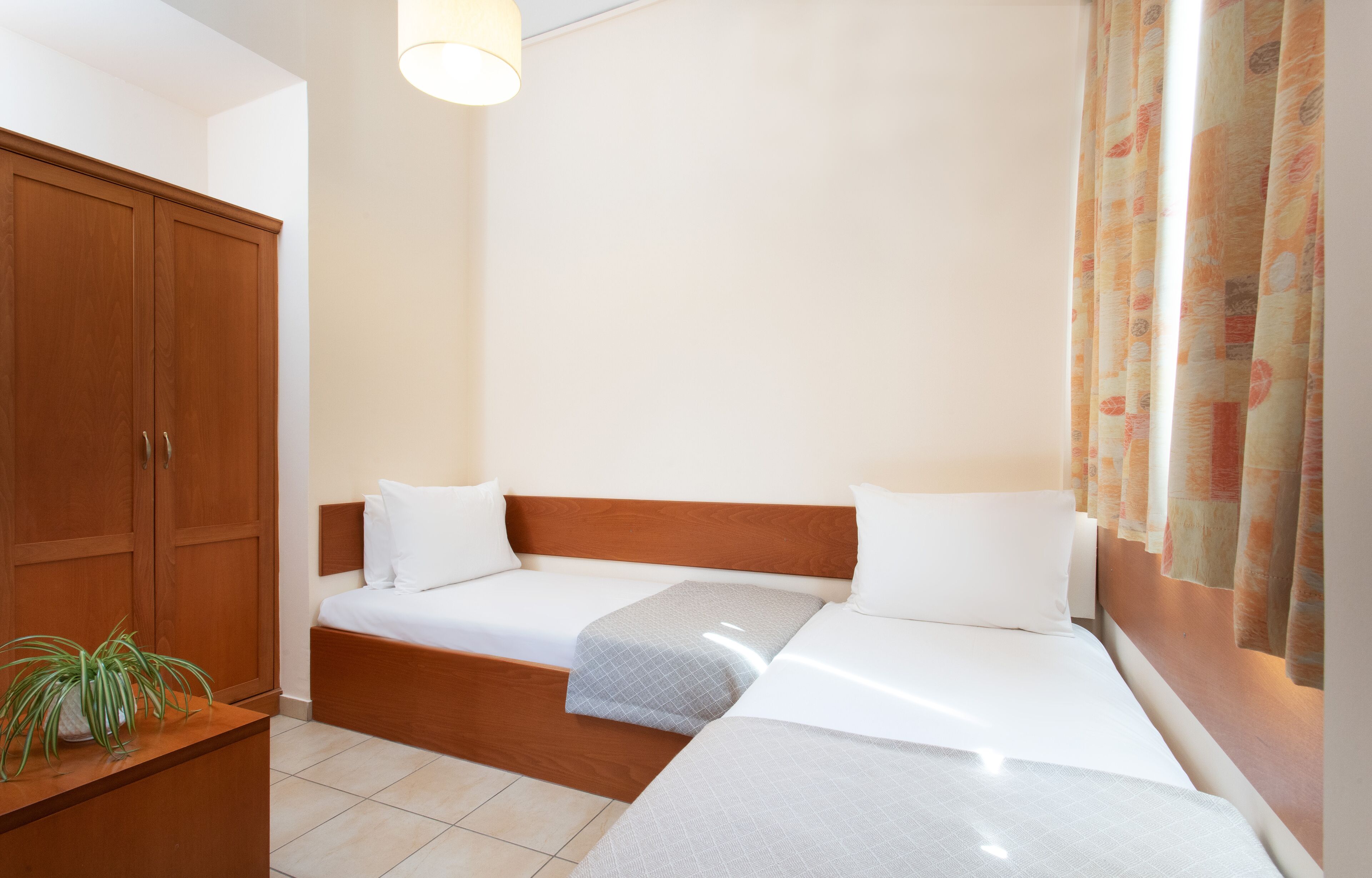 A room with two single beds, a wooden wardrobe, a bedside table with a plant, and a window with curtains.