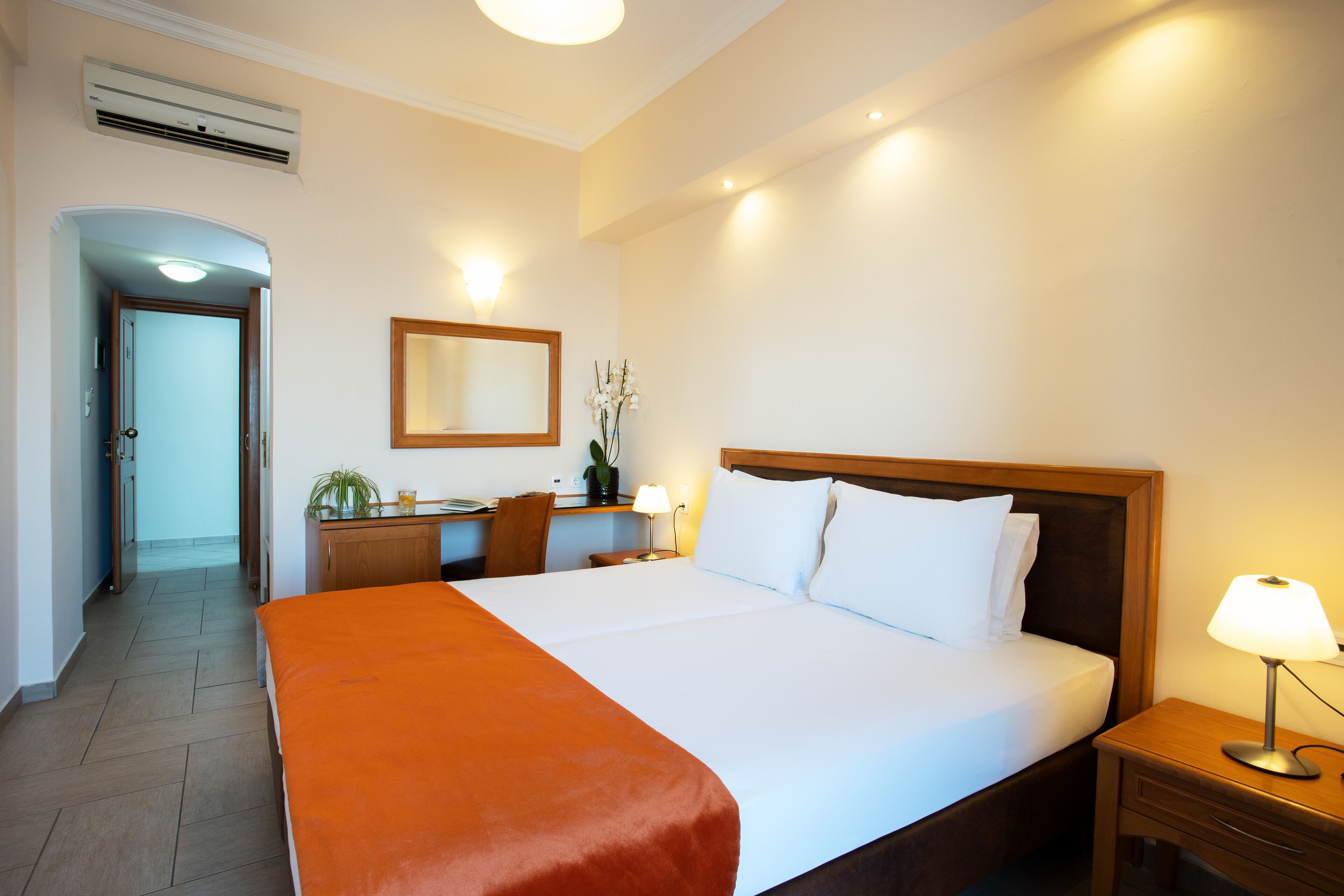 Una habitación de hotel con una cama grande, un escritorio con silla, un espejo y una mesita de noche con lámpara.