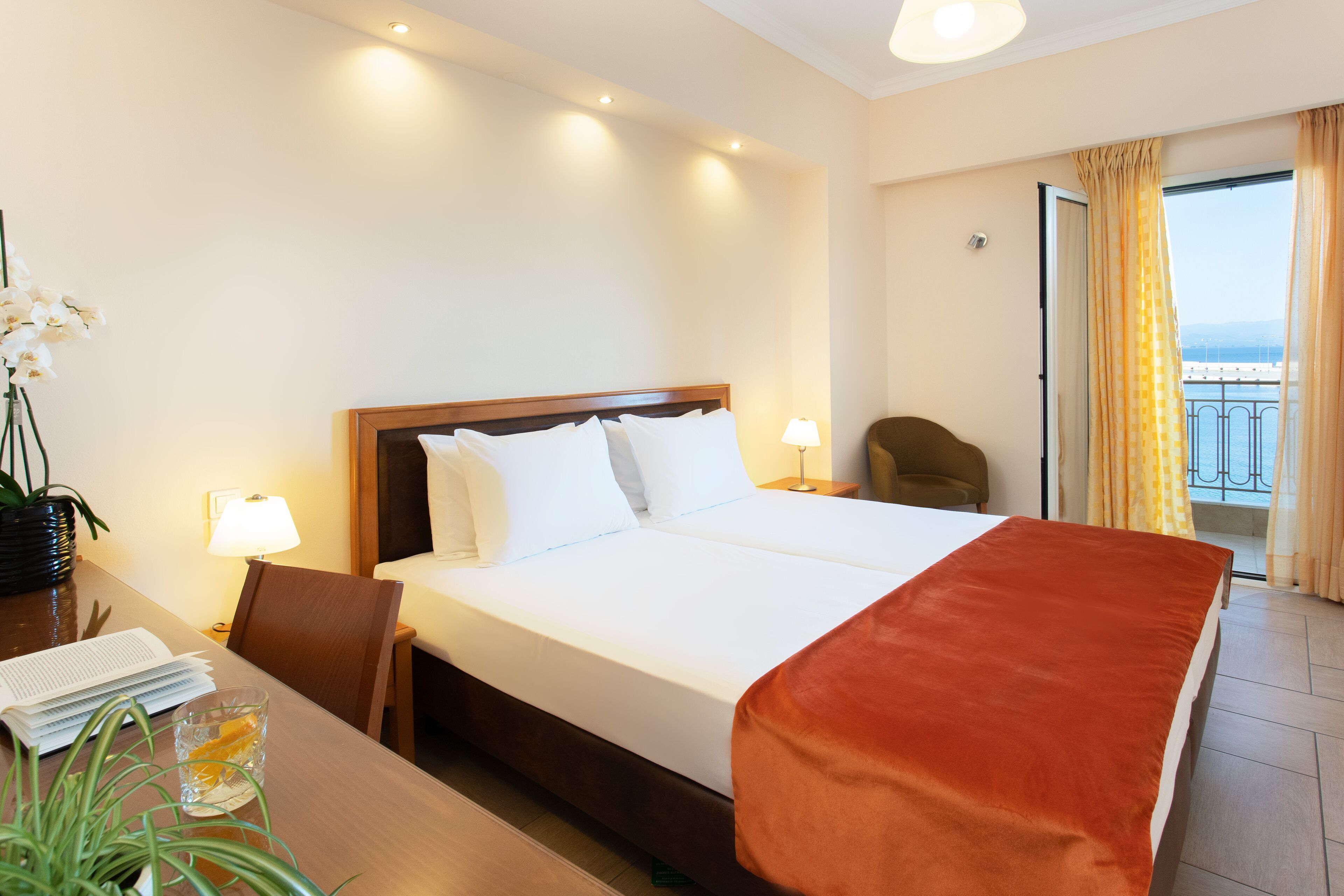 Ein Hotelzimmer mit einem großen Bett, einem Schreibtisch mit Lampe, einem Sessel und Meerblick vom Balkon.
