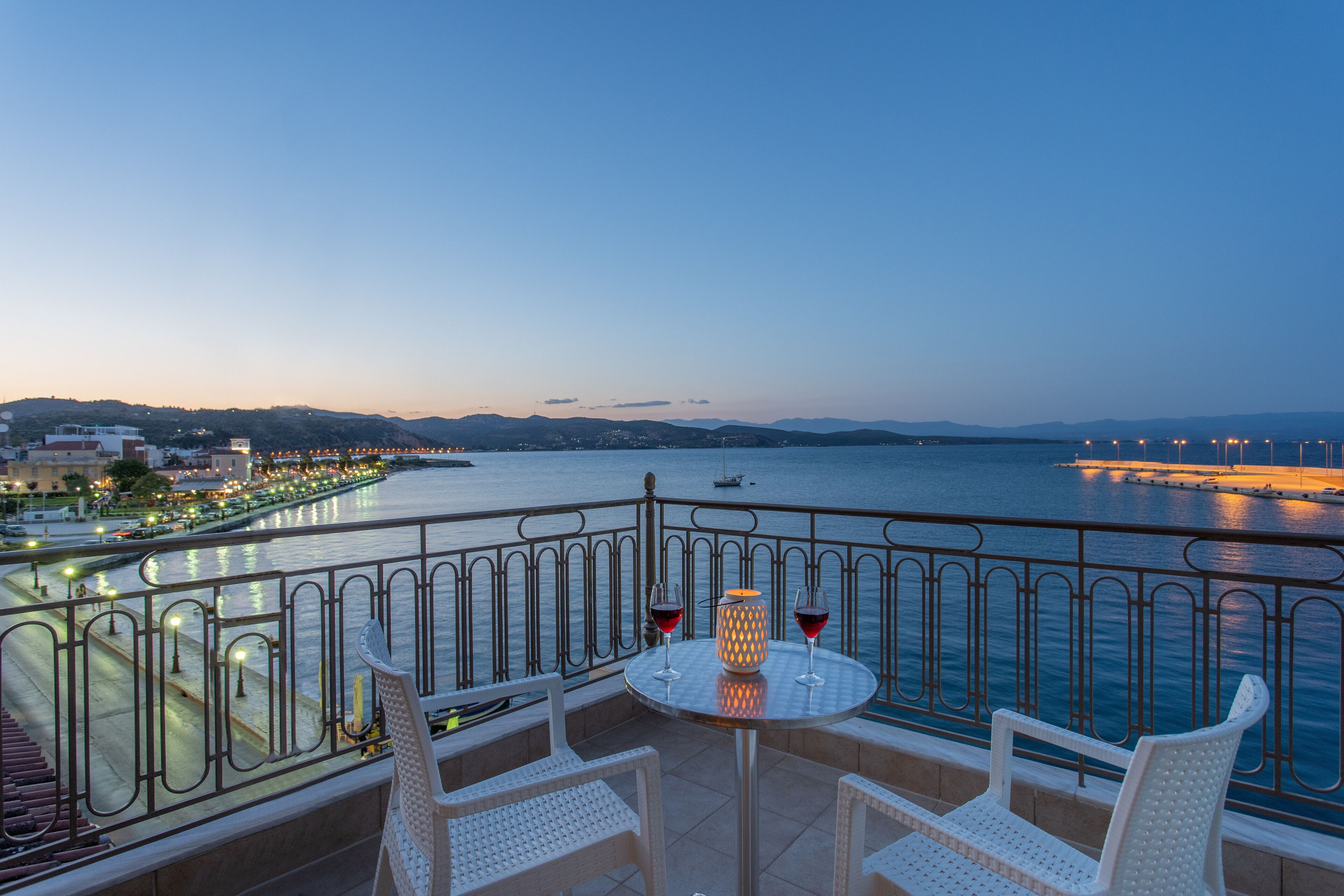 A balcony with a table and chairs overlooking a waterfront, with a lamp and two glasses of red wine.