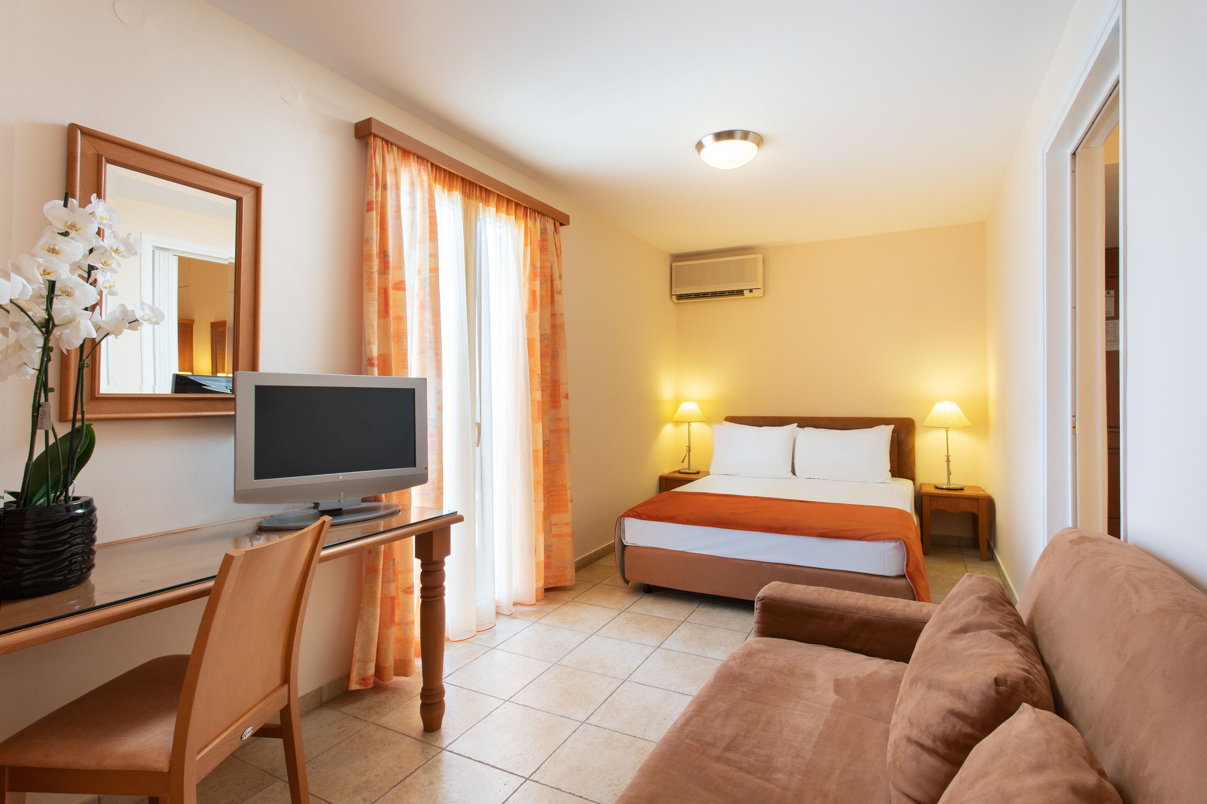 Ein Hotelzimmer mit einem Bett, einem Schreibtisch mit Computer, einem Fernseher, einer Couch und einer Topfpflanze.