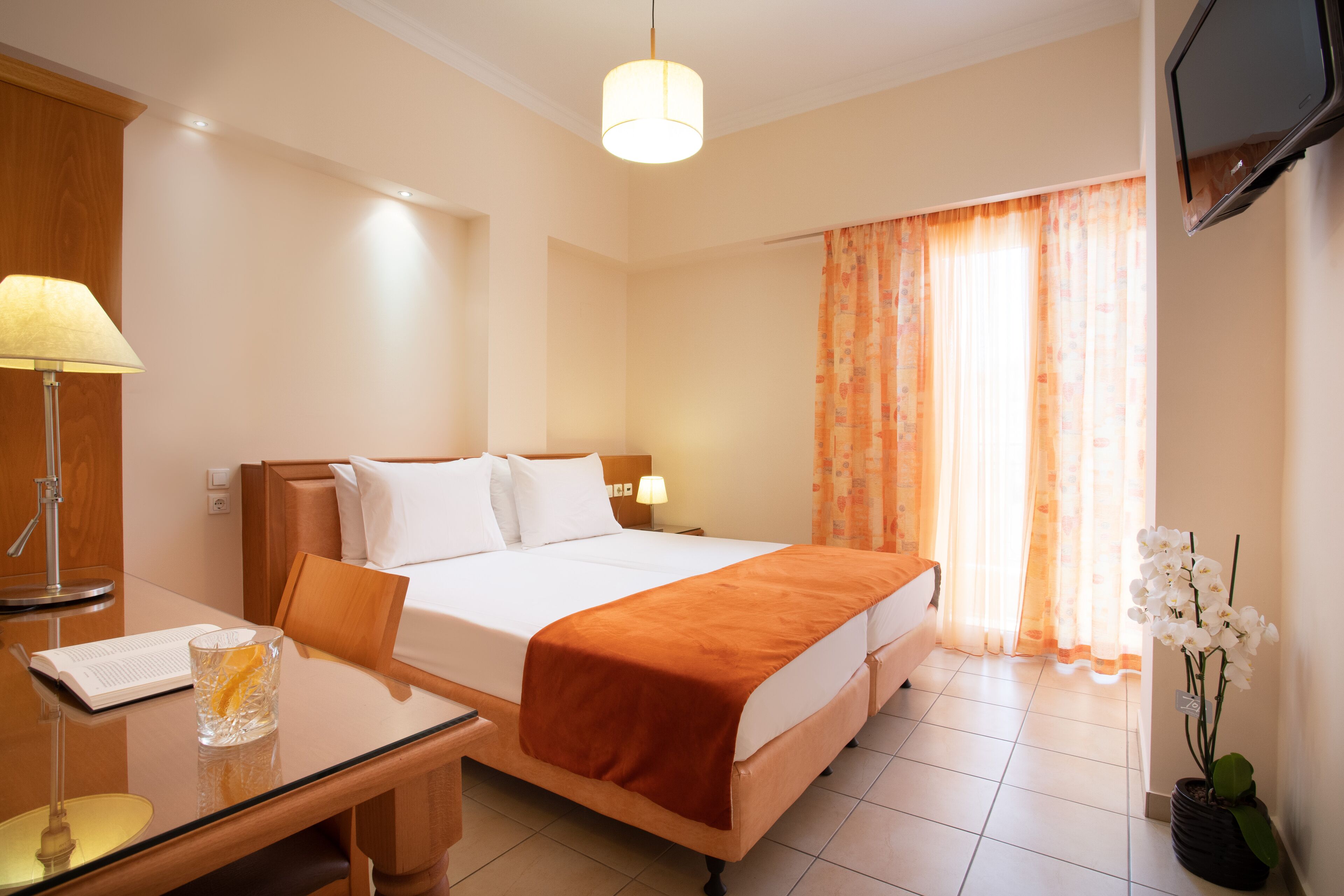 Una camera d'albergo con un letto, una scrivania con lampada, una TV e una finestra con tende.