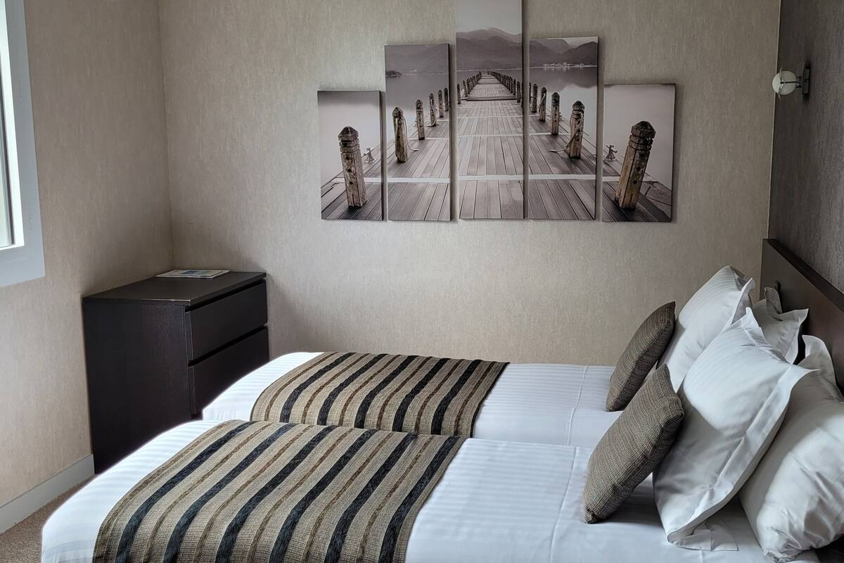 A neatly arranged hotel room with a bed, a bedside table, a radiator, a window, and a wall-mounted artwork depicting a pier.