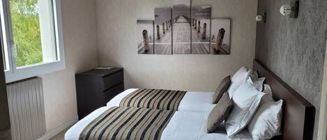 Ein ordentlich eingerichtetes Hotelzimmer mit Bett, Nachttisch, Heizkörper, Fenster und einem Wandbild, das einen Steg zeigt.