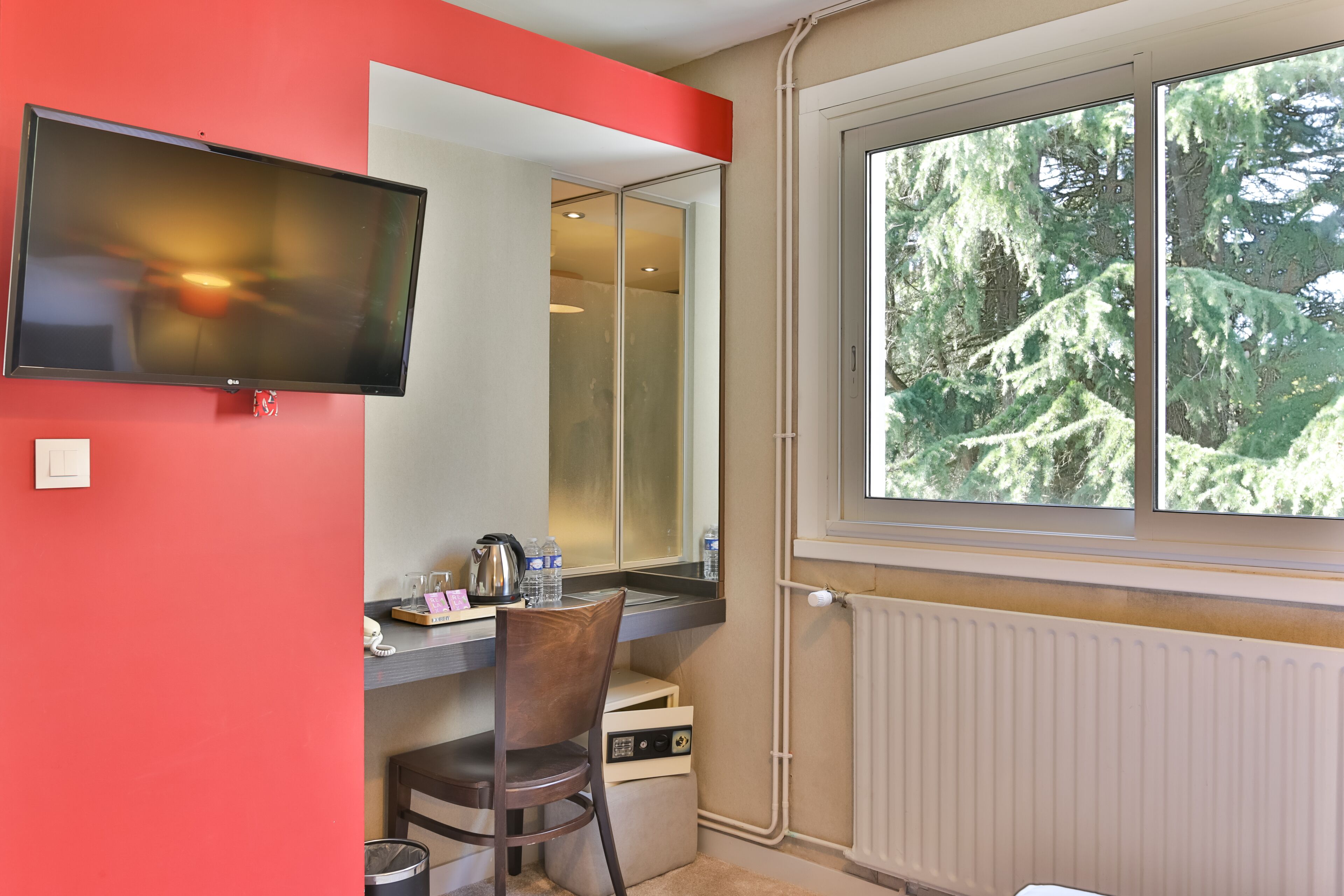 A room with a TV mounted on the wall, a window with a view of trees, a desk with a chair, and a radiator.