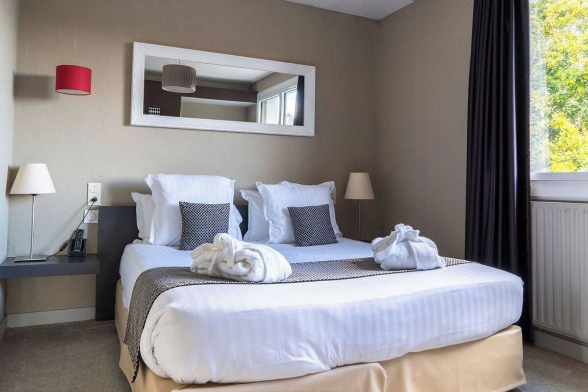 A hotel room with a bed, two bedside lamps, a mirror, and a window with curtains.