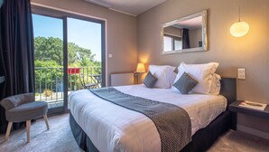 Habitación de hotel con una cama grande, una silla, un balcón con vista a árboles y un espejo en la pared.