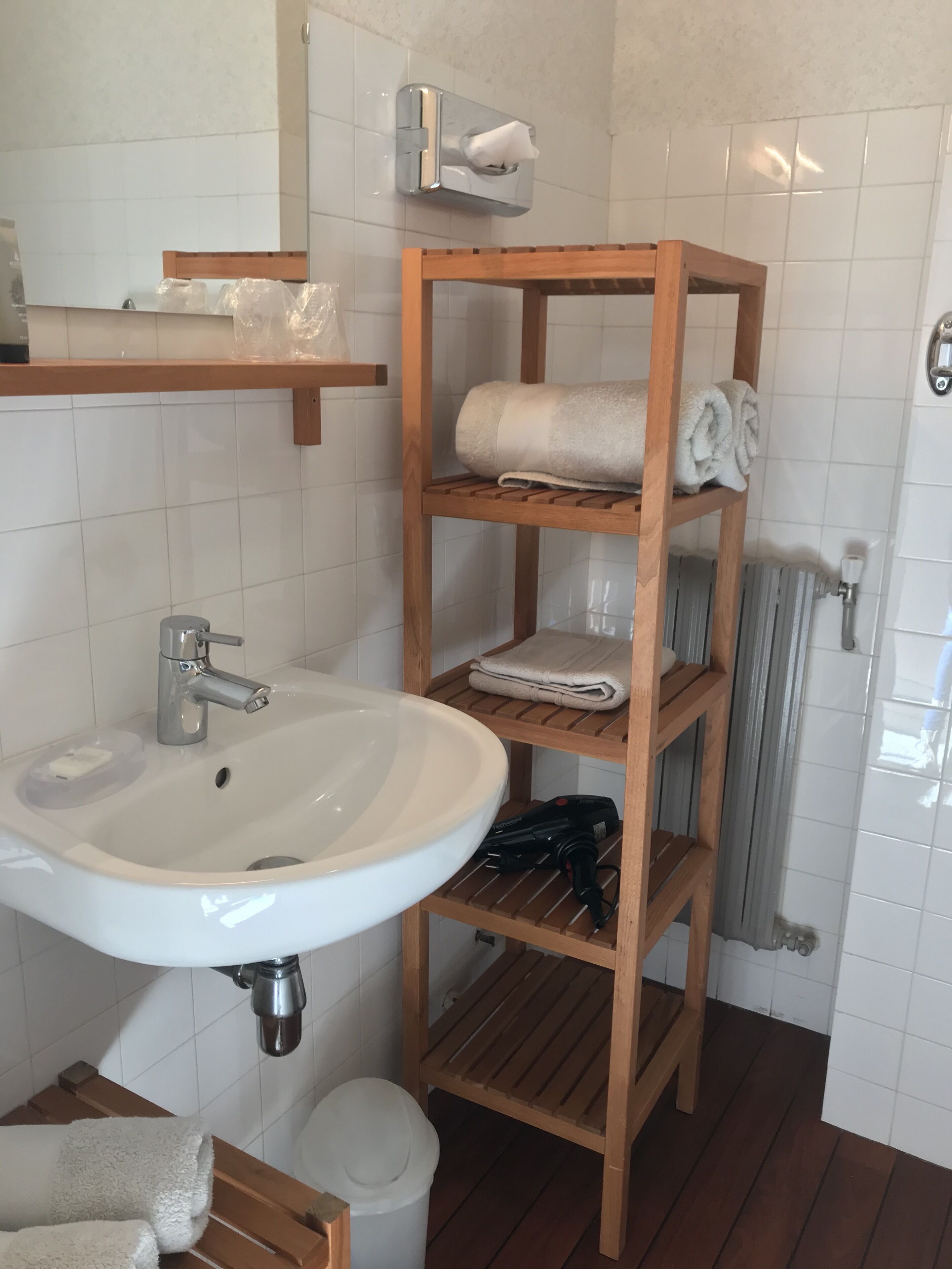 Une salle de bain avec un lavabo blanc, des étagères en bois où sont posées des serviettes, un sèche-mains et un radiateur.