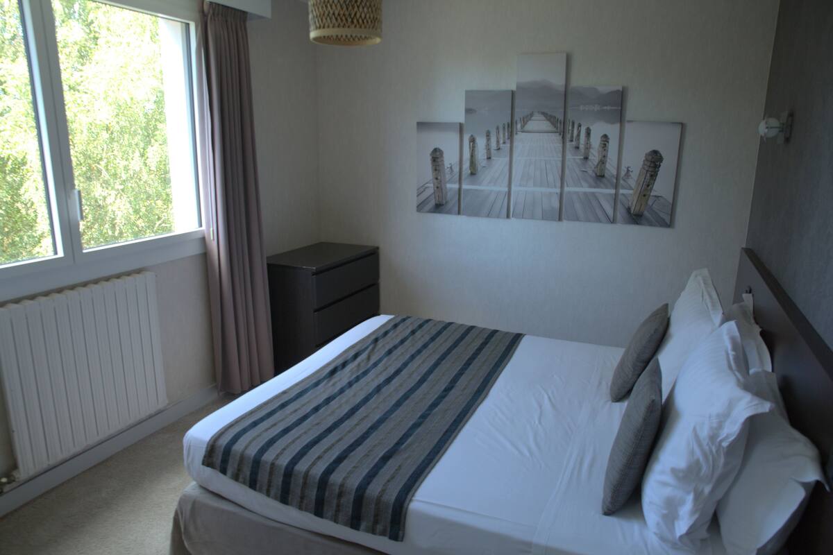 A neatly made bed with a striped blanket, a bedside table, a lamp, and a wall art depicting a pier.