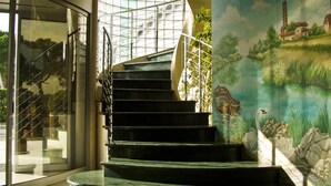 Un escalier avec une structure de verre menant à un palier, lequel est entouré d’une fresque représentant un paysage avec une église, une tour et des oiseaux.