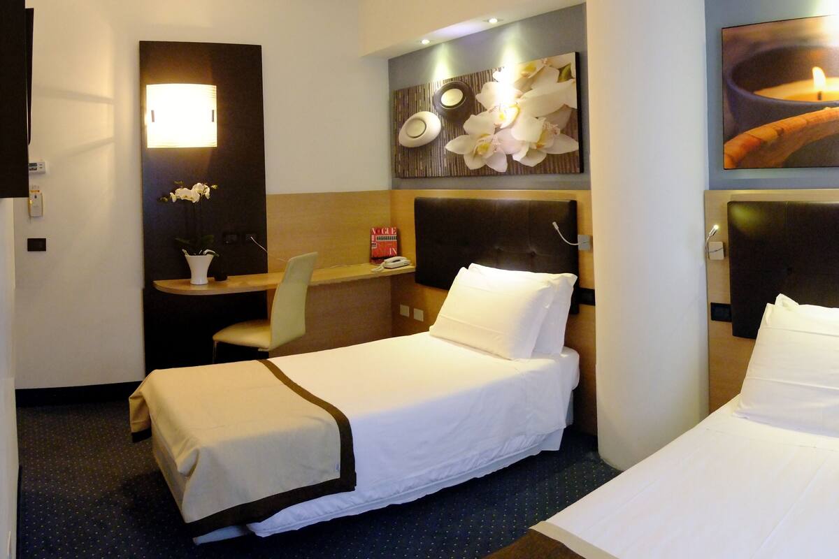 A hotel room with two single beds, a desk, a chair, a nightstand, and a wall-mounted TV.