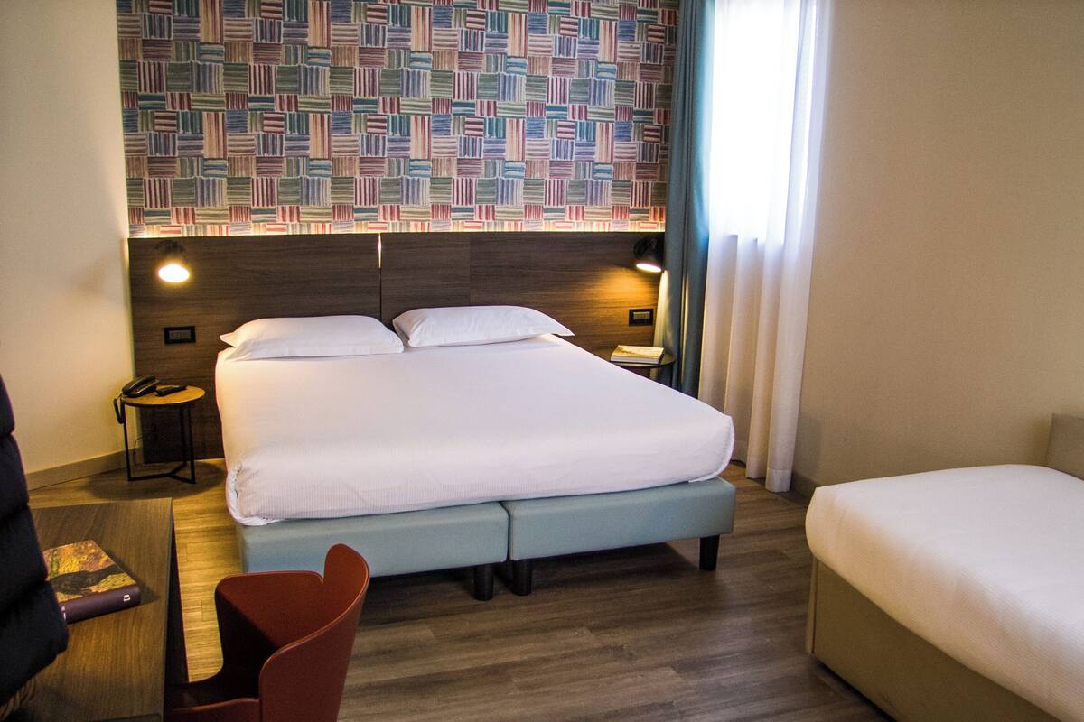 A hotel room with a large bed, a bedside table with a lamp, a chair, and a small desk with a telephone.
