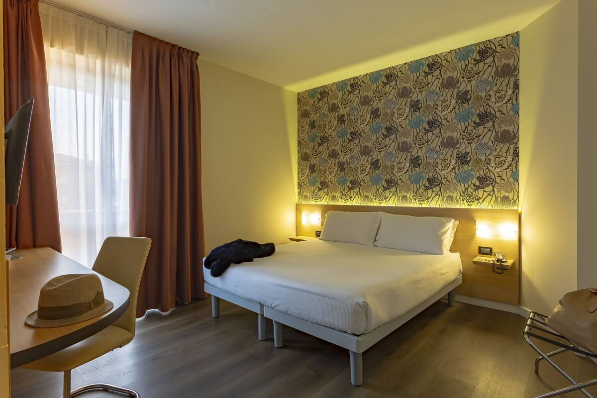 A hotel room with a bed, bedside table, chair, and a wall with a floral pattern.