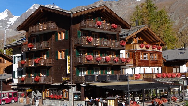 A multi-story wooden hotel with balconies adorned with red flowers, a sign indicating it's a hotel, and a view of snow-capped mountains in the background. - Hotel Tenne (Saas-Fee)