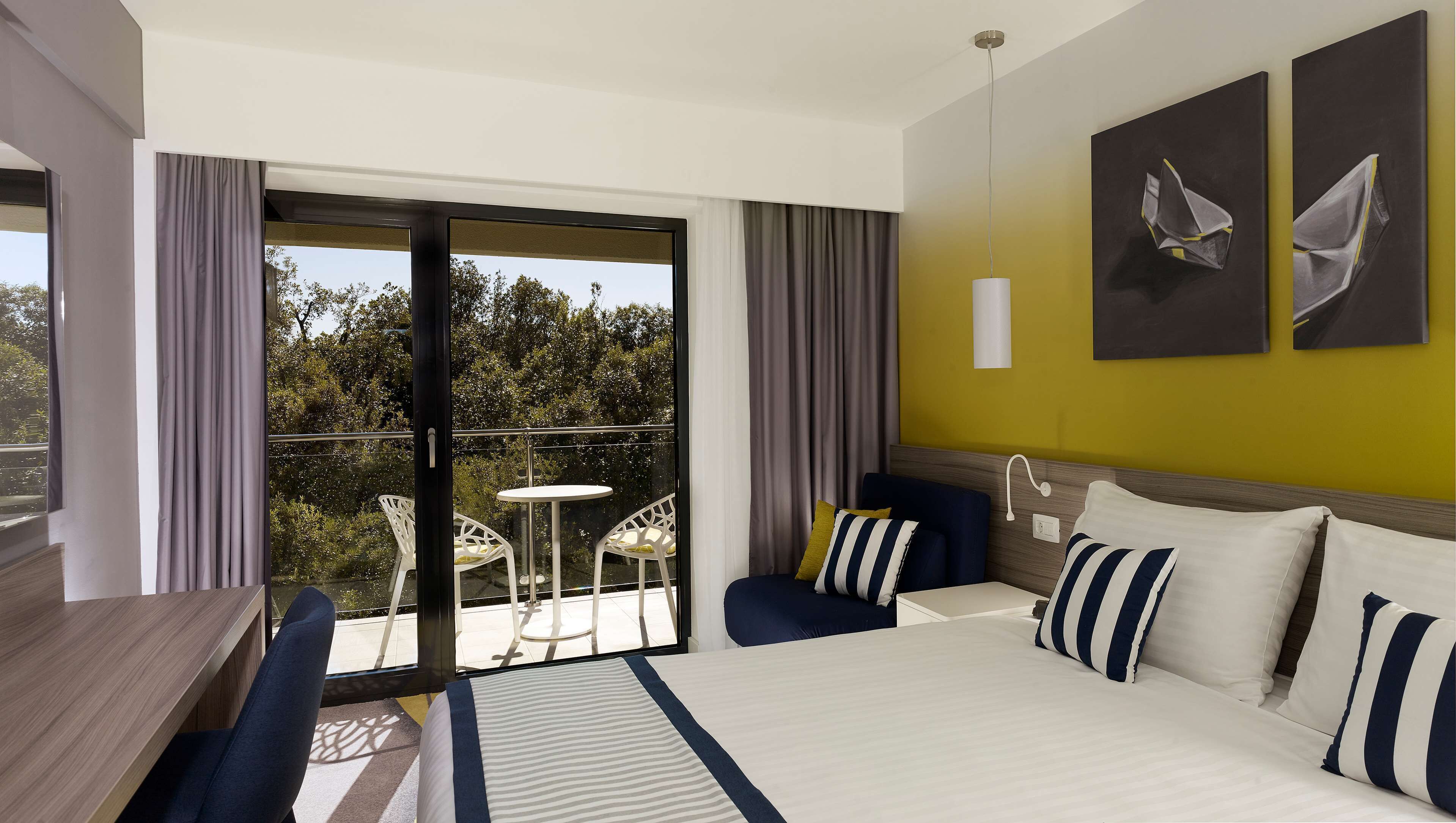 A hotel room with a bed, a desk, a chair, a balcony with a table and chairs, and two paintings on the wall.