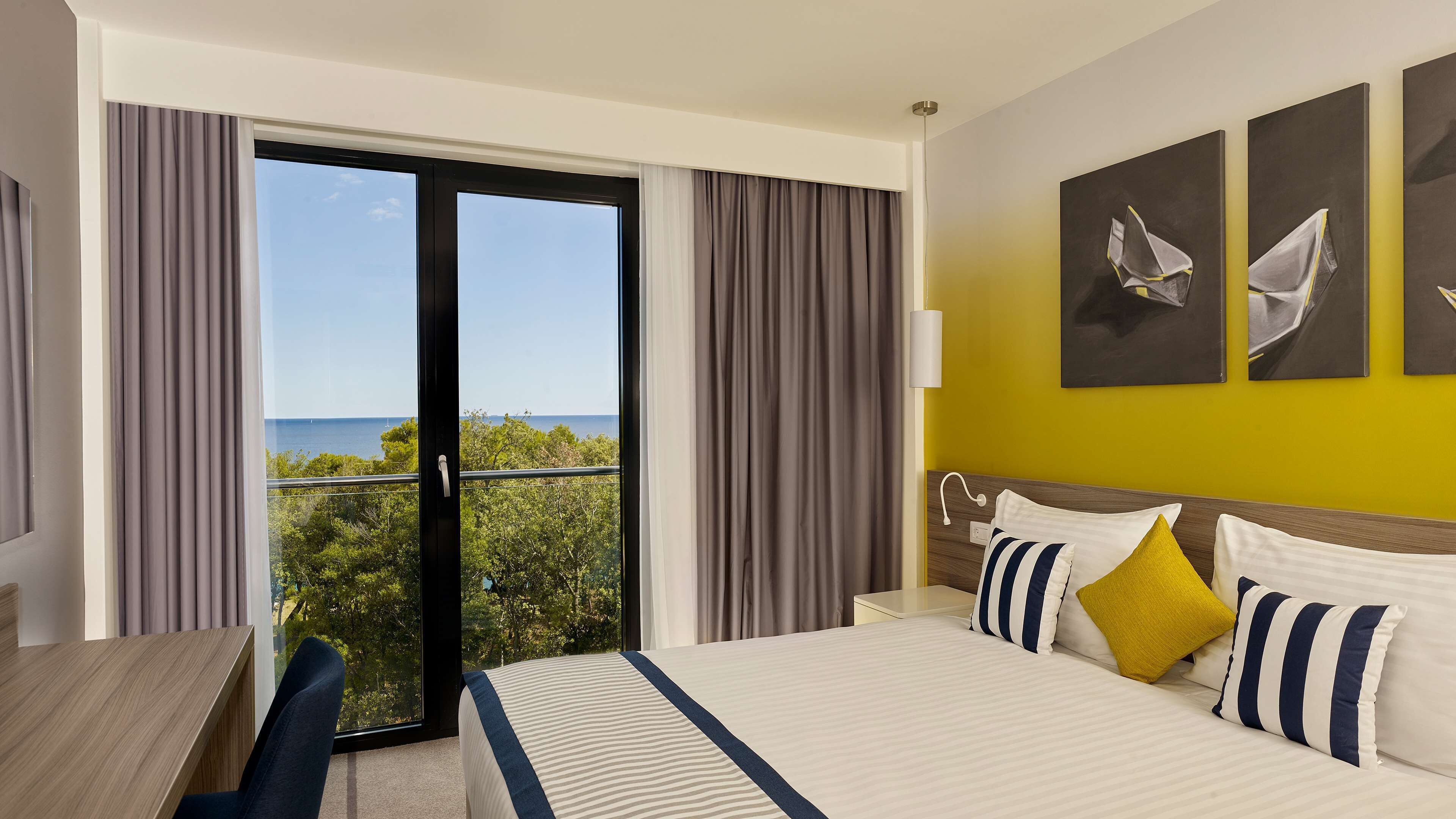 A hotel room with a bed, a desk, and a view of the sea through a sliding door.