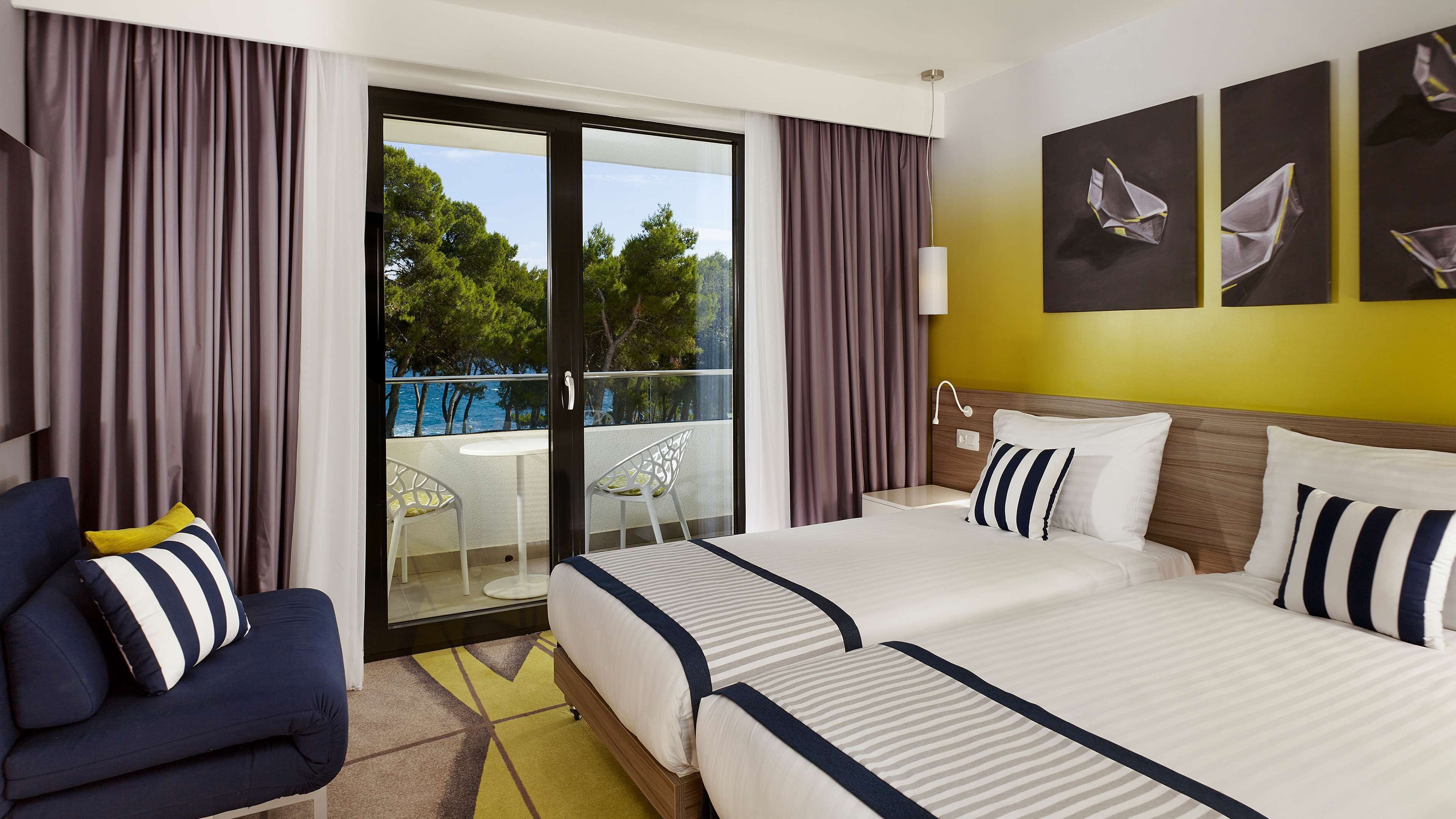 A hotel room with a large bed, a chair, a balcony with a view of trees and a body of water, and artwork on the wall.