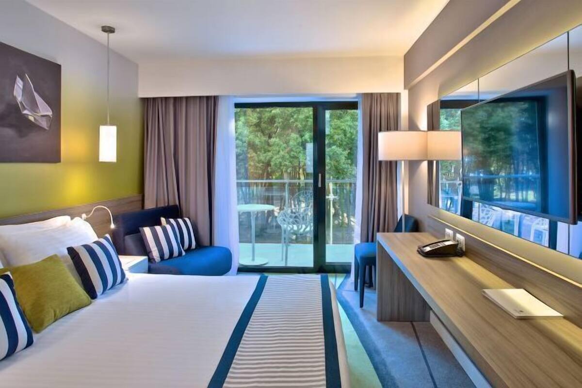 Quarto de hotel com cama, escrivaninha, cadeira, televisão e varanda com vista para árvores.