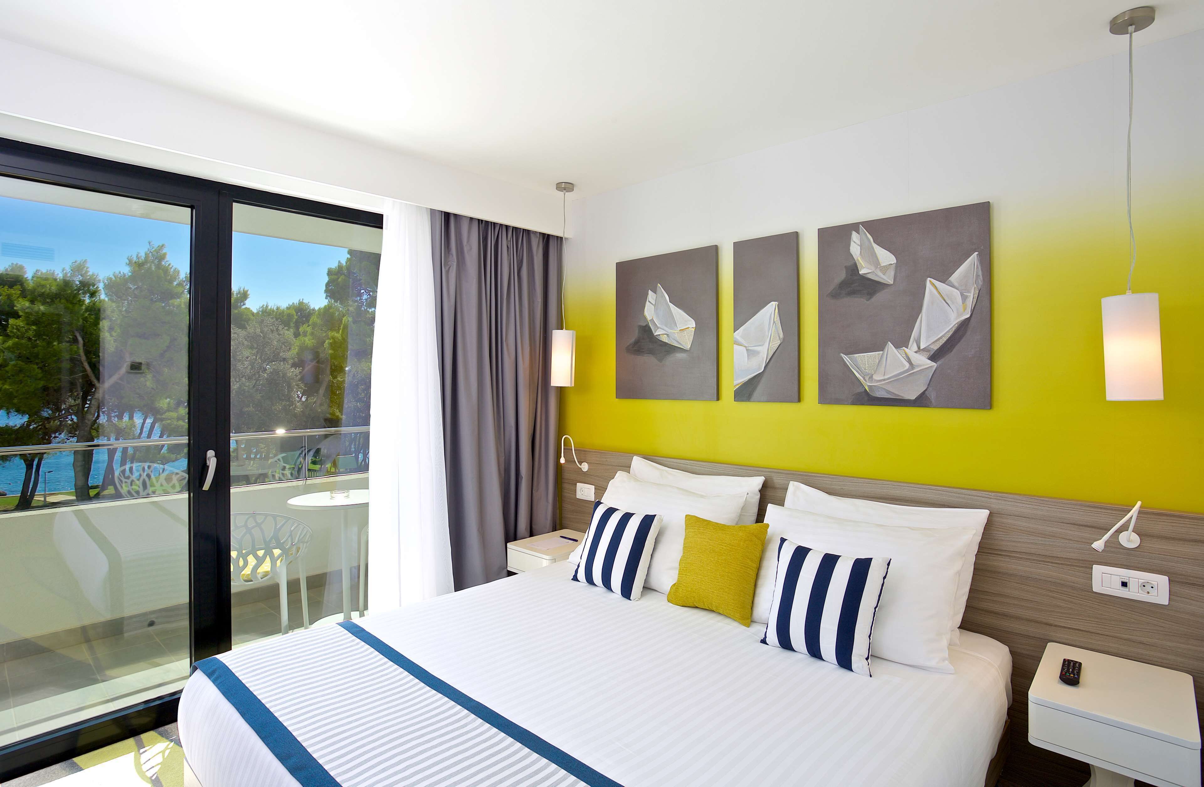 A hotel room with a large bed, a balcony with a view of trees and a body of water, and artwork on the wall.