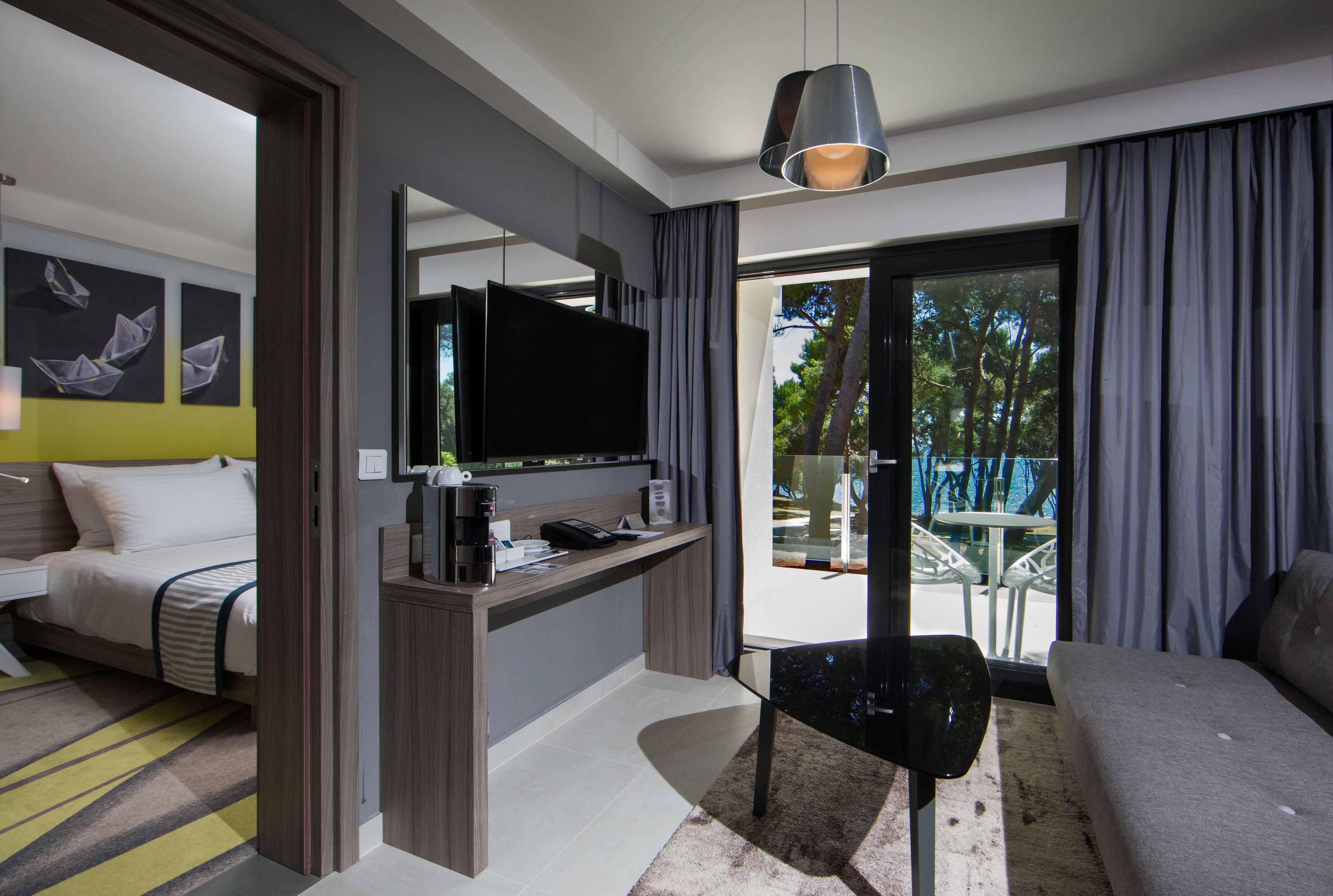 A modern hotel room with a bed, a desk with a flat-screen TV, a sofa, and a balcony with a glass door.